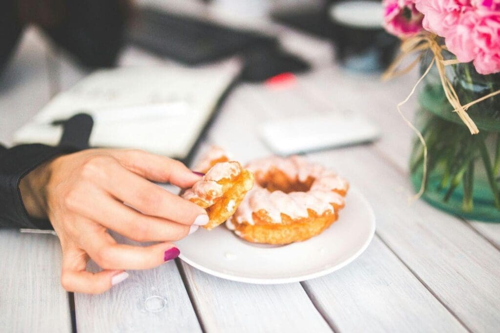 A person picking up a piece of a glazed donut from a white plate, representing emotional eating and food guilt