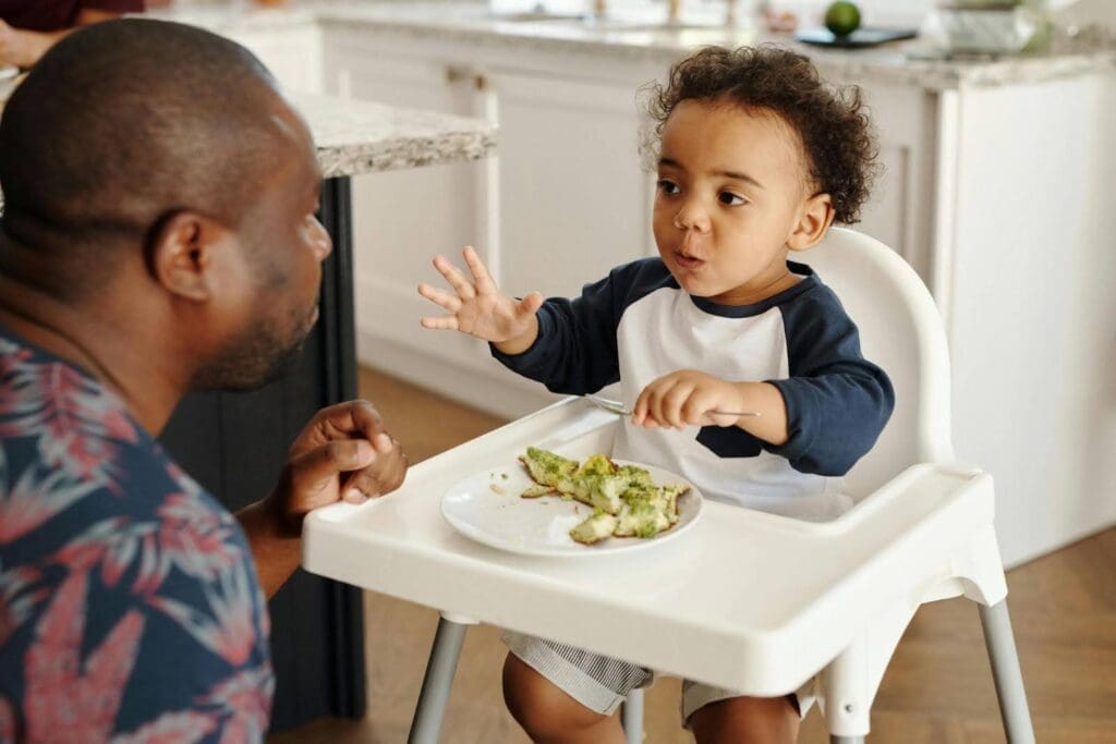 Toddler eating broccoli in a high chair while interacting with a parent, demonstrating picky eating strategies for a selective eater.