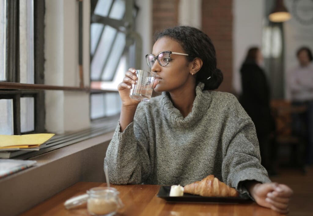 Woman sitting alone at a café table with a croissant, looking out the window — representing food anxiety and disordered eating