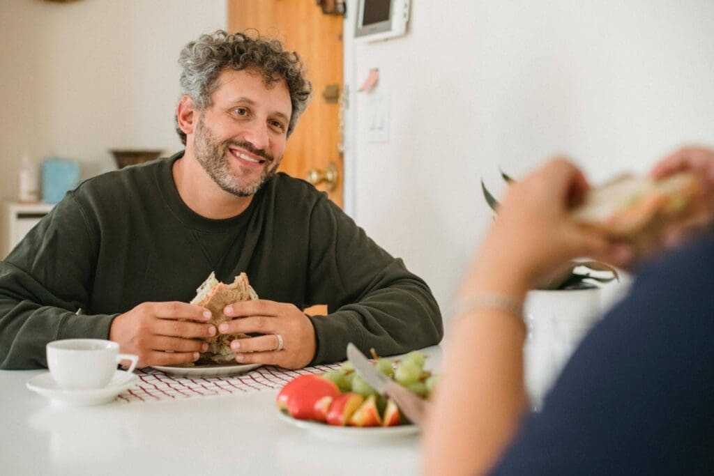 Man smiling while enjoying a bite of food at an outdoor table, illustrating a peaceful relationship with food and body trust.