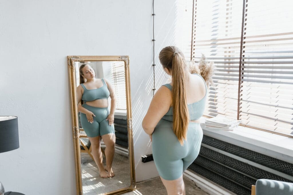 A plus-size woman in a green two-piece set looks at her reflection in a full-length mirror, illustrating a journey through body confidence issues, negative body image, and finding body positivity.