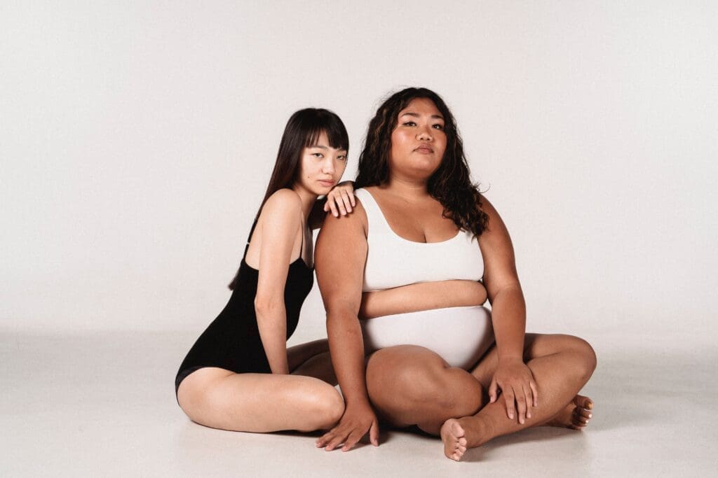 Two women of different body types sitting together in simple undergarments, representing body positivity vs body neutrality and advocating for a positive body image across all shapes.