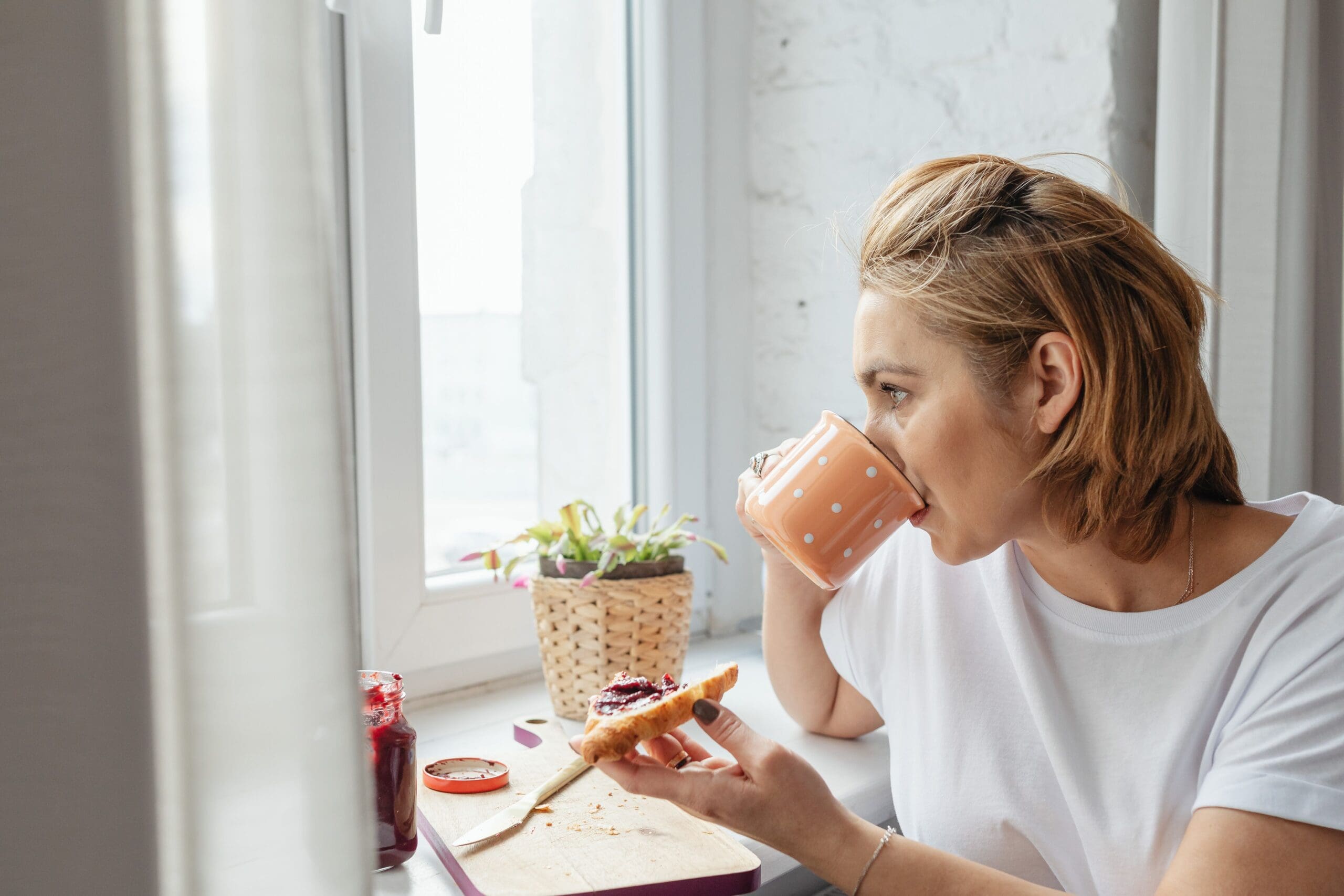 Woman calmly eating toast and drinking coffee by a window, modeling a peaceful relationship with food